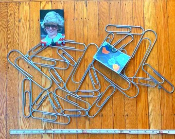 May include: A collection of large silver paper clips scattered on a wooden surface. A small photograph of a woman with sunglasses and a colorful top is among the clips. A small card with a floral design is also present. A ruler is at the bottom.