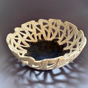 May include: A ceramic bowl featuring a cream-colored, lattice-style openwork rim with a textured, speckled surface. The interior base of the bowl is finished in a deep, dark blue glaze that contrasts with the light, porous-looking exterior structure.
