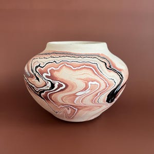May include: A ceramic vase with a wide, rounded body and a cream-colored rim. The vase features a swirling pattern of black, white, and shades of brown. The design gives the vase a marbled appearance, suitable for home decor.