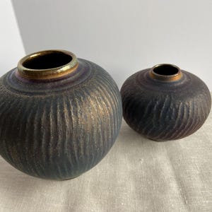 May include: Two ceramic vases with a textured, ribbed design. The vases are dark blue with gold rims. The larger vase is on the left, and the smaller vase is on the right. The vases are decorative.