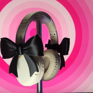 May include: White headphones with black satin bows on each earcup. The headphones are on a stand against a pink and white background.