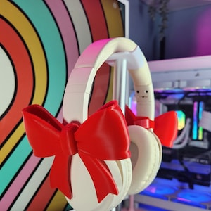 May include: Cream-coloured headphones with bright red bow accents. The headphones are displayed on a silver stand, with a colourful striped background. The earcups are round and the headband is padded.