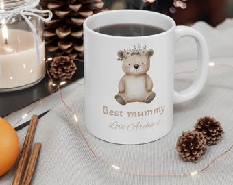 Può includere: Tazza in ceramica bianca con un orso marrone che indossa una corona di fiori. La tazza ha il testo "Best mummy Love Archie x" stampato su di essa.