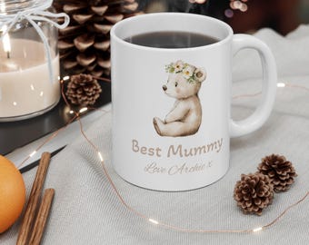 Può includere: Tazza in ceramica bianca con un orso marrone che indossa una corona di fiori. La tazza ha il testo "Best Mummy Love Archie x" stampato su di essa.