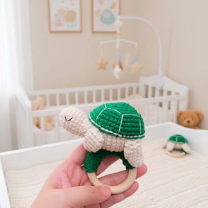 May include: A handmade crochet turtle baby rattle. The rattle features a green shell, beige head and limbs, and a wooden ring. The turtle is held in a hand, with a crib and mobile in the background.