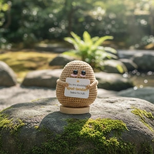 Puede incluir: Una figura de patata tejida a ganchillo con una cara sonriente y brazos pequeños, sosteniendo una tarjeta que dice "You are absolutely spud-tacular! Believe this truth." La figura es de color beige con ojos negros y nariz rosa, sobre una base de madera.