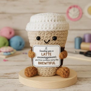 May include: A crocheted coffee cup with a smiling face and a white lid. The cup holds a sign that reads "Sending you a LATTE encourggement; you brew excellence every day BREWTIFUL." The cup is brown, beige, and white, and sits on a wooden base.