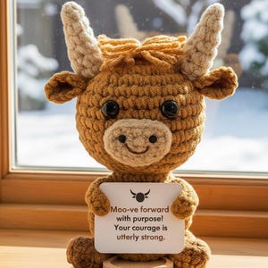May include: A crocheted brown bull figurine with large black eyes and tan horns. The bull holds a sign that reads "Moo-ve forward with purpose! Your courage is utterly strong."