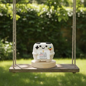 May include: A white crocheted controller-shaped plush toy with black and colorful button details, holding a sign that reads "Never hit the PAUSE button on your dreams. Keep leveling up!" The toy is on a wooden base, suspended on a swing.