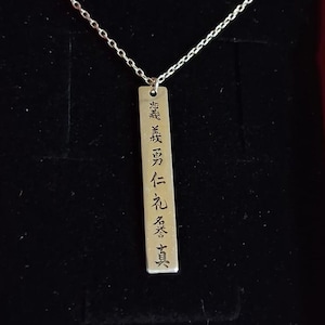 May include: Silver-toned necklace with a rectangular pendant. The pendant features engraved Japanese characters. The chain is delicate and the pendant hangs against a dark background. The necklace is a piece of jewelry.