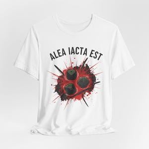 Puede incluir: Camiseta blanca con un diseño gráfico. El diseño presenta tres dados negros y la frase latina "ALEA IACTA EST" en texto negro, sobre un fondo rojo y negro salpicado.