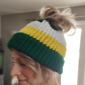 May include: A green, yellow, and white striped crocheted beanie with a messy bun hole in the top.