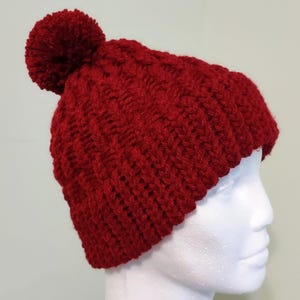 May include: A knitted red beanie hat with a matching pom-pom on top. The hat is made of a thick, textured yarn and is designed to keep the wearer warm. The hat is shown on a white mannequin head.
