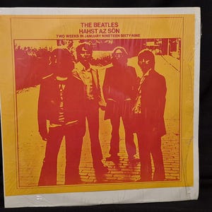 May include: A square vinyl record cover featuring a red-toned monochromatic photograph of The Beatles standing outdoors. The text reads THE BEATLES HAHST AZ SÖN TWO WEEKS IN JANUARY NINETEEN SIXTY-NINE. The background is a solid yellow border.