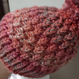 May include: A hand-knitted hat in shades of coral, pink, and gray. The hat features a textured, woven pattern and a fluffy pom-pom on top. The hat is displayed on a white mannequin head.