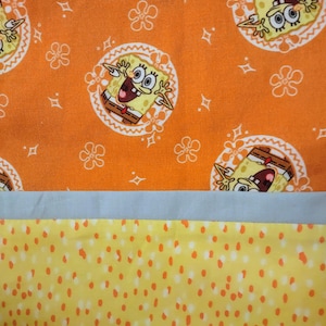 May include: Orange fabric with a repeating pattern of SpongeBob SquarePants in white circles, surrounded by white floral and diamond shapes. Below is a light blue strip, followed by yellow fabric with orange polka dots. The fabric is likely for a craft project.
