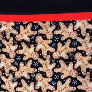 May include: A fabric with a repeating pattern of gingerbread men with red bow ties and snowflakes on a black background. A red stripe separates the patterned fabric from a solid black top.