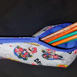 May include: A pencil case with a blue zipper, filled with colored pencils. The case is white with a cartoon print of Mario Kart characters. The interior is blue.