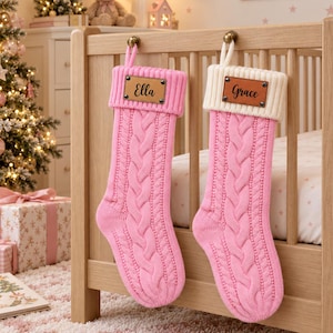 Op de afbeelding: Twee gebreide kerstsokken met kabelpatroon hangen aan een houten ledikant. De linker sok is effen roze met een roze boord en een leren label met de naam Ella. De rechter sok is roze met een crèmekleurige boord en de naam Grace.