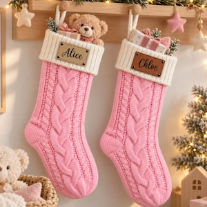 Op de afbeelding: Twee roze kerstsokken met kabelpatroon en witte geribbelde boorden. Elke sok heeft een rechthoekig bruin label in lederlook met de namen Alice en Chloe in zwarte letters gegraveerd. De sokken zijn gevuld met kleine cadeautjes.