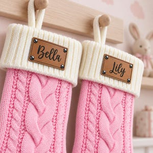 May include: Two pink cable-knit Christmas stockings with cream-coloured ribbed cuffs and hanging loops. Each stocking features a rectangular tan leather-like patch attached with four metal rivets, displaying the names Bella and Lily in black script.