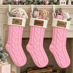 May include: Three pink cable-knit Christmas stockings with white ribbed cuffs. Each stocking features a brown leather-look name tag labelled Lily, Mabel, or Amelia. The stockings are filled with plush toys, including a bunny, bear, and unicorn.