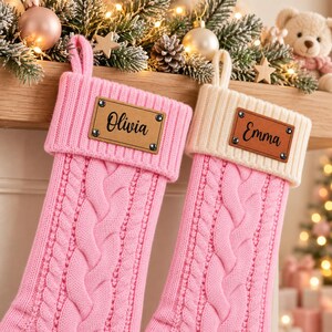 Puede incluir: Dos calcetines navideños de punto trenzado con cuerpo rosa y puño doblado. El calcetín izquierdo tiene un puño rosa con una etiqueta de cuero marrón con el nombre Olivia. El derecho tiene un puño crema con una etiqueta de cuero con Emma.