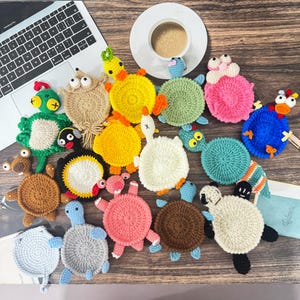 Peut inclure: Une collection de dessous de verre ronds et colorés au crochet représentant divers animaux, notamment des tortues, des canards et d'autres créatures avec des corps en laine texturée, de petites têtes et des membres, disposés sur une surface en bois.