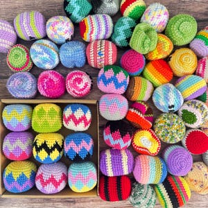 May include: A collection of small, round, crocheted balls in various bright colors and patterns, including zigzags, stripes, and solid hues. Some balls are arranged inside a square cardboard box, while others are scattered around the exterior.