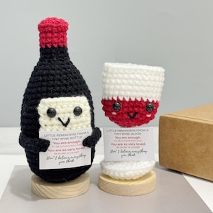 May include: A pair of crocheted wine-themed items: a bottle and a glass. The bottle is black with a red top, the glass is red and white. Both have smiling faces and hold cards with text.