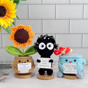 Op de afbeelding: Drie handgemaakte gehaakte figuren: een zonnebloem in een pot, een zwart figuur en een blauwe vuilnisbelt. Elk houdt een bord met tekst vast. De zonnebloempot leest "Emotional Support Plant". Het zwarte figuur en de vuilnisbelt zeggen "It's fine."