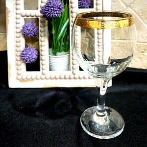 May include: A clear glass goblet featuring a wide, rounded bowl and a sturdy stem. The rim of the glass is adorned with a thick, metallic gold band that displays an intricate, repeating decorative pattern around its entire circumference.