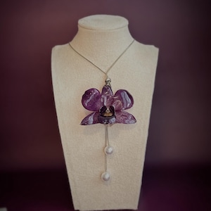 Peut inclure: Un collier pendentif orchidée violette avec deux perles blanches pendantes. Le pendentif en forme de fleur présente des pétales violets foncés et une chaîne argentée. Le collier est présenté sur un buste de mannequin beige sur fond violet foncé.