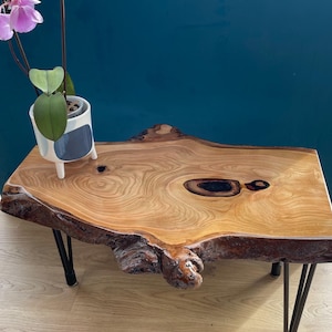 Może przedstawiać: Stolik kawowy z naturalną krawędzią, jasnobrązowym blatem z drewna i ciemnobrązowymi krawędziami. Stół ma czarne nogi typu hairpin. Na stole stoi mała biało-niebieska doniczka z fioletową orchideą.
