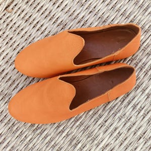 Puede incluir: Un par de zapatos sin cordones de cuero naranja. Los zapatos tienen una puntera redondeada y un perfil bajo, con un acabado liso y mate. El interior es de color marrón contrastante.
