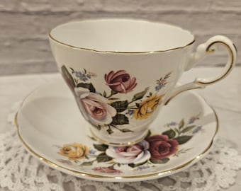 Può includere: Una tazza da tè e un piattino bianchi con un motivo floreale con rose rosa, gialle e viola. La tazza e il piattino hanno un bordo dorato.