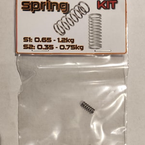 Peut inclure: Un sac en plastique transparent contenant deux ressorts argentés. Le sac porte une étiquette indiquant "SELIGARMOR Spring KIT" et "S1: 0,65 - 1,2 kg S2: 0,35 - 0,75 kg".