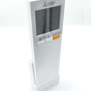 Peut inclure: Une télécommande blanche Mitsubishi Electric pour climatiseur avec un écran noir. La télécommande comporte des boutons étiquetés TEMP, +, et OFF/ON. La télécommande est dans un support blanc.
