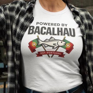 Könnte beinhalten: Ein weißes T-Shirt mit dem Text "POWERED BY BACALHAU" und einer Fischgrafik in den Farben der portugiesischen Flagge. Die Person trägt ein schwarz-weiß kariertes Hemd, blaue Jeans und einen Schweißhelm.