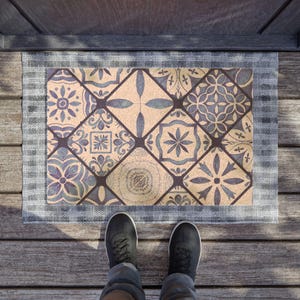 May include: A rectangular doormat with a geometric tile pattern in shades of blue and beige. The mat is framed by a gray border. Black shoes and blue jeans are visible in the foreground, standing on a wooden surface.