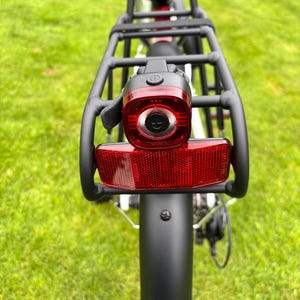 Könnte beinhalten: Rückansicht eines Fahrrads mit schwarzem Gepäckträger und rotem Rücklicht. Das Rücklicht ist rechteckig mit kreisförmiger Mitte und schwarzem Gehäuse. Das Fahrrad hat einen schwarzen Kotflügel und steht auf einer grünen Rasenfläche.