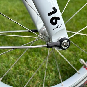 Könnte beinhalten: Nahaufnahme eines Fahrradrads mit einer weißen Gabel. Auf der Gabel steht in schwarzer Schrift "16 INCH". Das Rad hat silberne Speichen und einen schwarzen Reifen. Der Hintergrund ist grünes Gras.