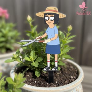 May include: A decorative garden stake featuring a cartoon character with black hair, glasses, and a straw hat, wearing a light blue shirt and dark blue skirt. The character holds silver garden shears and stands on a black base in soil.