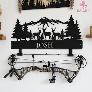 May include: Black metal wall art featuring a mountain range, trees, and deer silhouettes, with the name "JOSH" below. A compound bow is mounted below the art. The bow has a camouflage pattern.