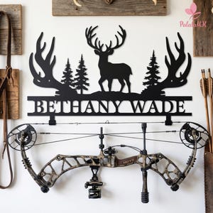 May include: Black metal deer silhouette wall art with the name "BETHANY WADE" below. A camouflage compound bow hangs below the sign. The background includes wooden panels and hunting-themed decor.