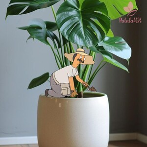 May include: A decorative plant stake featuring a cartoon character wearing a wide-brimmed tan hat, a white shirt, and gray pants, kneeling while holding a small trowel. The character is positioned in a large, smooth, light-colored ceramic planter.