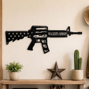 May include: A black metal wall art piece shaped like a rifle, featuring a cutout American flag pattern with stars on the stock and the text WALKER'S ARMORY engraved on the handguard. The design includes a detailed barrel, sight, and magazine.