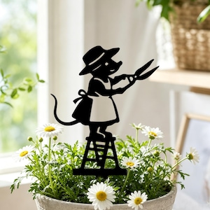 May include: A black metal garden stake silhouette featuring a mouse wearing a hat and apron, standing on a small ladder while holding a pair of garden shears. The decorative figure is positioned upright within a potted plant with white flowers.