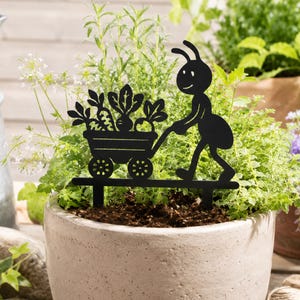 May include: A black metal garden stake featuring a silhouette of an ant pushing a small cart filled with various vegetables. The design includes two pointed prongs at the base for insertion into soil, showcasing a flat, cutout artistic style.