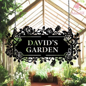 Puede incluir: Letrero de jardín de metal negro con el texto "DAVID'S GARDEN". El letrero está decorado con rosas, hojas y recortes de mariposas. Cuelga de cadenas frente a un invernadero lleno de plantas.